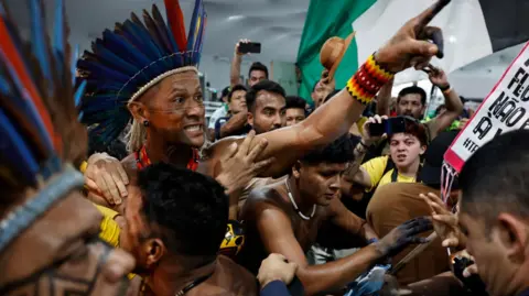 A protester, wearing a traditional headdress, raising his hand at a protest. 