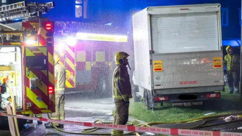 Melksham News A fire officer in yellow fire officer clothing looking at the scene with a 'fire do not cross' tape in the foreground of the picture.
There are two fire vehicles behind him and another fire officer sorting a hose. 
Although the photo is taken at night, the picture is lit up by floodlights from the fire appliance. 