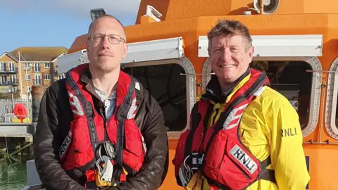 Chris Haffenden, wearing a red RNLI lifejacket, stands beside RNLI volunteer Andy, who is wearing a yellow RNLI jacket. They are both on the deck of an orange lifeboat moored at a harbour. Buildings and railings are visible in the background under a bright sky.