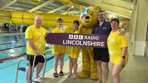 BBC Five BBC Lincoln colleagues smiling for the camera in front of a pool all wearing bright yellow tops with Pudsey's face on along with Pudsey ears and a Pudsey teddy in the middle. Holding up a purple BBC Radio Lincolnshire sign. Pudsey himself is in the photo - a yellow bear with a colourful spotted eye patch over one eye. 