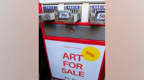 A red, white and silver vending machine with a sign which reads 'Art For Sale'. There is a yellow sticker which says '50p'.