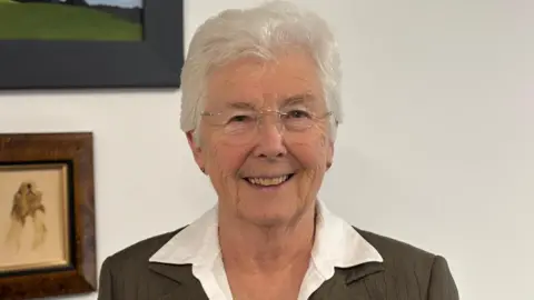 LDRS A lady with short white hair, unframed glasses, a white shirt and brown-grey jacket smiles at the camera