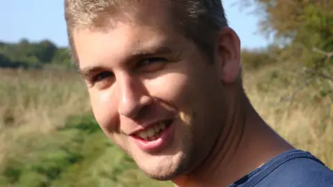 A photo of Alex Young in a field on a sunny day. He is smiling at the camera and has short blonde hair and wears a blue T-shirt. 