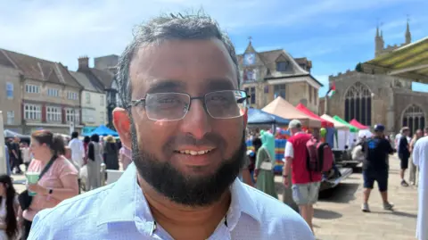 A head and shoulders image of a man wearing glasses and a light coloured shirt. He is standing in a city centre square and looking into the camera smiling. Behind him are stalls and people walking around. 