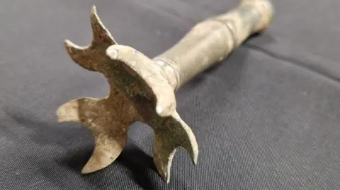 The rattle is made of metal and appears heavily weathered, with visible corrosion, tarnish, and oxidation. It consists of a long cylindrical shaft or handle attached to a decorative, multi-lobed head.