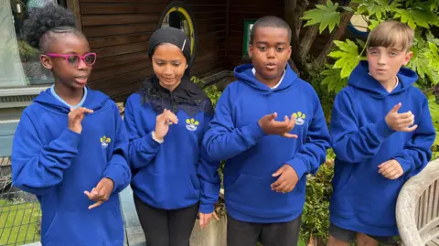 Ollie Conopo/BBC Four pupils, outside, in a school, by plants, all doing sign language. They have their right arms out. 