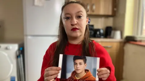 Heidi Rose Elliott, wearing a red top. She has red lipstick on and brown shoulder length hair, and is photographed in her kitchen. She is holding an AI-generated photo of her son