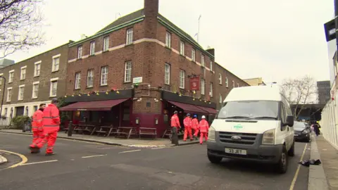 Construction workers wearing orange fluorescent clothing walking past pub on corner of a road junction with a van parked on the road