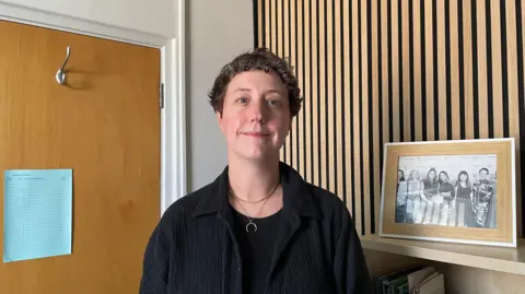 BBC/Sofía Luis-Hobbs A woman with short, dark hair smiles at the camera. She wears a black top under a black shirt worn open around two gold necklaces. There is a black and white group photo on a shelf beside her.