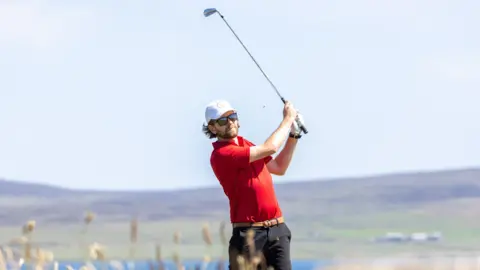 Vannin Photos Daryl Callister wears a red T-shirt and dark trousers, with a white cap and dark sunglasses. He swings a silver golf club high over his shoulder with water and low hills in the background.