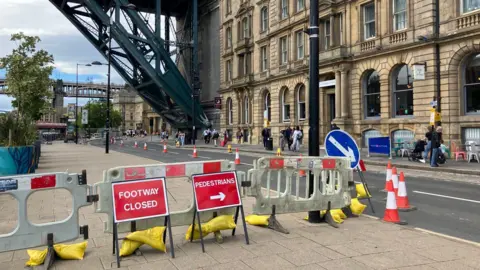 A red footway closed sign and pedestrian sign in front of plastic barriers blocking the path underneath the Tyne Bridge. Traffic cones are lining the middle of the road.