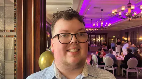 Oisin Ager is standing in an events space with different tables set out and a blue projector screen. He has dark glasses and a light coloured shirt. He has brown hair.
