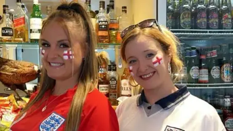 Two women in England football tops with England facepaint stand in front of a fully-stocked bar smiling