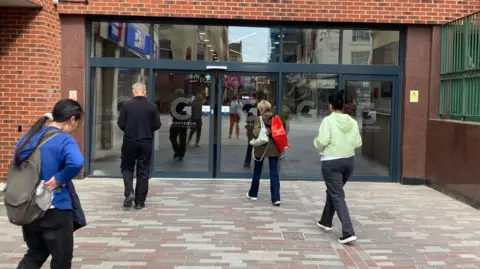 Alex Pope/BBC Four people walking towards the entrance of a shopping centre, which has large glass doors with a grey frame. There is block paving in front of the entrance. All four people have their backs to the camera.
