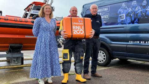 A lifeboat coxswain in uniform holds an orange box containing the scroll alongside a woman in a blue dress and a man in dark trousers  