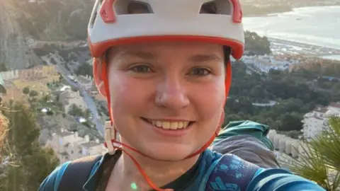 Aimee O'Doherty Aimee O'Doherty, a woman wearing a white helmet with orange straps, is taking a selfie whilst climbing. Behind her is a seaside town, a considerable distance beneath her. She is smiling for the photo.