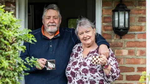 Lol and Sue Hepworth are standing outside their home and they are both holding a mug each. Lol is wearing a navy jumper and Sue is wearing a flowery shirt.