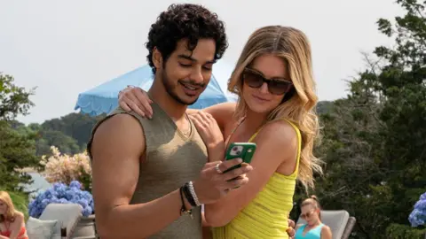 Netflix Uma cena do programa The Perfect Couple da Netflix com o personagem Shooter de Ishaan vestindo um colete marrom claro ou verde e segurando um telefone enquanto a personagem Meritt, vestindo uma blusa amarela, coloca o braço em volta dele. Há árvores verdes e paisagens ao fundo.