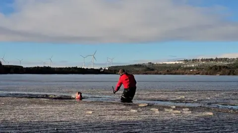 Frankie, with a swimming cap and goggles, swims through a channel in the icy Sweethope Lough. Coach Fenwick Ridely is standing in the water, knee deep, in a red coat and fluffy hat and monitoring her progress.