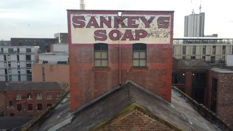 Jukebox PR The roof of Beehive Mill in Manchester, showing the Sankeys Soap red and white painted sign on a brick wall, with the city's skyline of flats and office buildings in the background