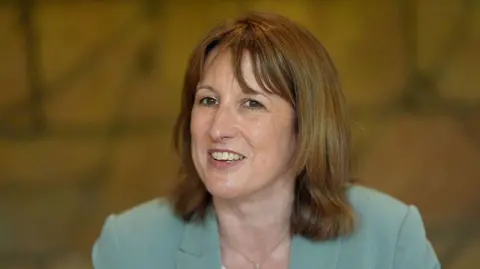 Getty Images A smiling Rachel Reeves wearing a sky blue jacket and gold necklace with a top of a white top just visible in the bottom of the frame. Behind her is a mottled yellowish brown background.