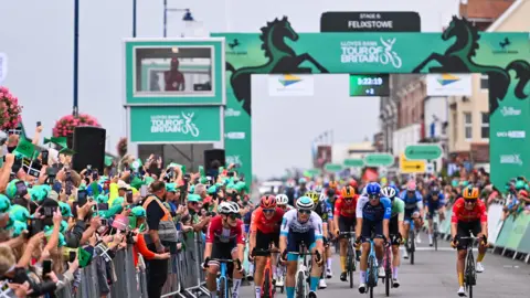 British Cycling Cyclists are coming towards the camera on a road. There are metal fences either side of the road where people are standing watching. A sign goes over the road which is green and has the horse logo for Lloyds. 