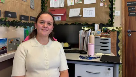 BBC Charlotte Moore is sitting at her desk in the classroom she teaches in. She is smiling and wearing a white top. Her desk has a computer monitor and there are various items pinned up on the wall.