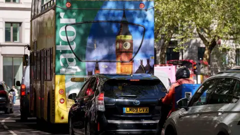 A rear view of a black Toyota Prius sitting in traffic on a city street, positioned directly behind an open-top double-decker tour bus featuring a large advertisement with an image of Big Ben and the text "Viator".