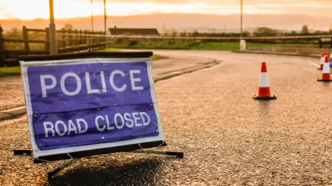 Getty Images A blue and white police sign on a road. Orange and white cones are also on the road. The background is the road and grass areas on either side. 