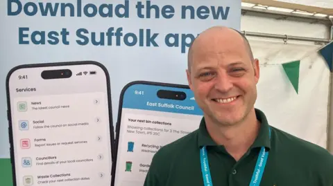 Vikki Irwin/BBC Ben Woolnough is standing in front of a poster for a new app which would help explain the new waste collection services. He is wearing a green polo shirt and is smiling at the camera. 