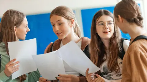 Getty Images Four teenage girls holding blank sheets of paper and discussing exam results