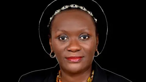 PA Media Lydia Mugambe is wearing a achromatic  chapeau  and metallic  concatenation  connected  her head, and earrings.