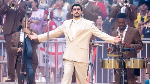 Bad Bunny in the centre with musicians surrounding him and a crowd in the background