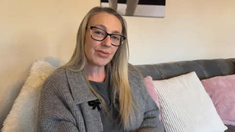 Alex Cooper-Kite sitting on a sofa. She is wearing a grey cardigan. She is wearing glasses and has long blonde hair. We can see white and pink pillows behind her and a picture hanging up on the wall. 