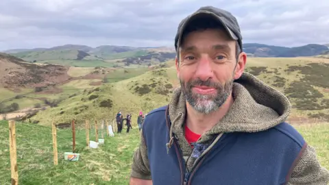A man in his 40's is looking into the camera. He's wearing a cap and has a beard. He's wearing a brown hoodie and a blue gilet. He's standing on a hill. In the backdrop there are a number of hills. 