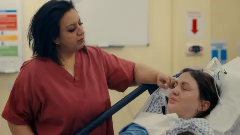 Channel 5 A woman in red scrubs wiping away a tear from the face of a woman who is laying in a hospital bed