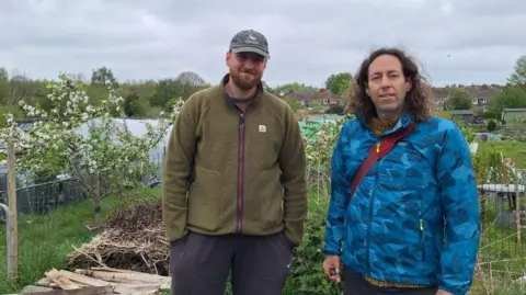 LDRS Volunteers James Pitter And Karl Scaplehorn stand looking at the camera in a set of allotments with some small trees behind them. A grey day.