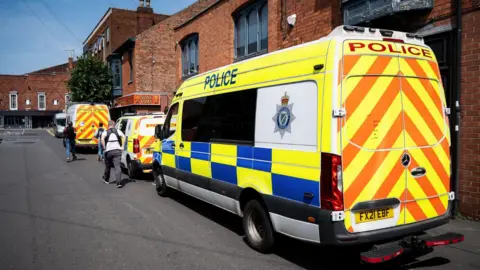 Three police vans parked on the right hand side of a road in front of a block of flats. Three people can be seen walking away from the camera. The sky is blue. 