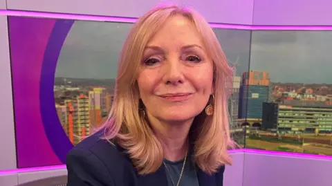 Image shows the Mayor wearing a black suit jacket. She has shoulder length blonde hair and is sitting in a BBC Radio Leeds studio, with an image of the city skyline behind her. 