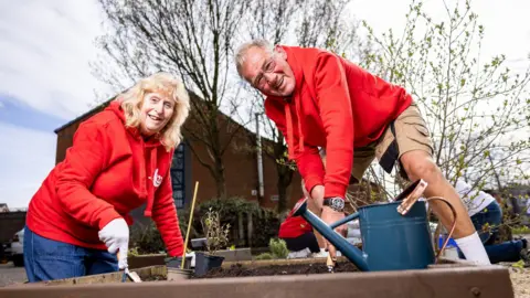 Allwyn Two people in red hoodies are gardening in raised beds. A man and a woman are smiling as they plant and water plants. 