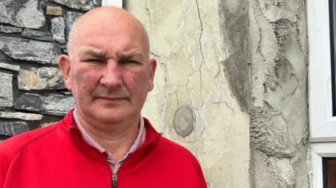 Ali Farren is wearing a red fleece and a white and red striped shirt. He is standing near a house with defective concrete blocks.