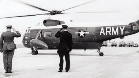 RAF Waddington Heritage Centre A black and white photograph of a US Sikorsky military helicopter about to take off. Two men stand nearby, both are saluting.