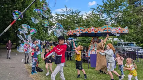 Provided Children are chasing and popping balloons at the festival. In the background is a merry-go-round.