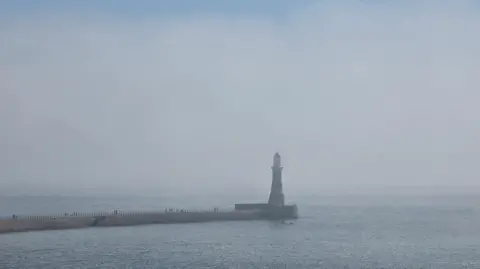Gill Helps The pier in Sunderland with the lighthouse on the end is covered in mist. The sea is mostly flat and the tone of the sky is a cold grey-blue.