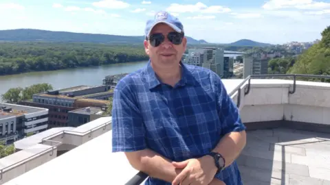 Contributed A man on a sunny day stands outside on a large balcony. He wears a blue cap and blue chequered shirt. There is a river behind him and several buildings.