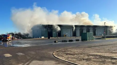 Cambridgeshire County Council Smoke rising from a large warehouse into a blue sky. On the left is a fire engine and a pile of waste. 