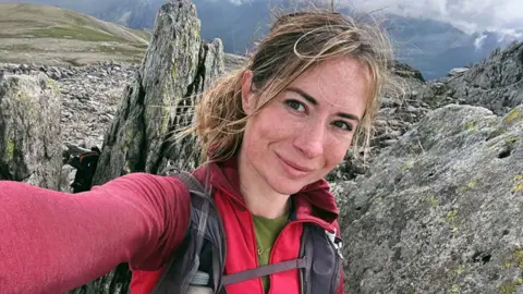Person taking a selfie on a rocky mountain ridge, wearing a red hiking top and backpack, with rugged stone formations, grassy slopes, and dramatic cloudy skies in the background.