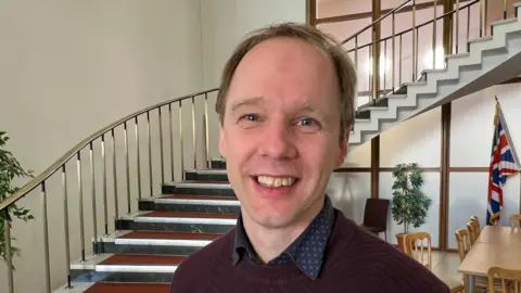 Councillor Jonathan Chilvers is wearing a maroon jumper over a dark shirt with white shapes on it. He is standing in a building lobby, with the Union flag showing in the right hand side of the picture
