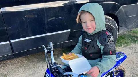 Nikita Burnett 4 year old in plastic upper-body armour and a sage green wind break. He is sat on a small blue moped and is eating chips out of a white take-away box
