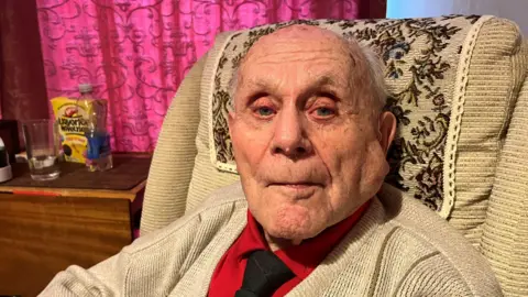 Stanley Eastgate, 102, has very thin grey hair on the sides of his head he has a red shirt with black tie under a beige cardigan and is sitting in a wing chair with a small table beside him with a glass and bottle of water on and some liquorice novelties in a yellow box.